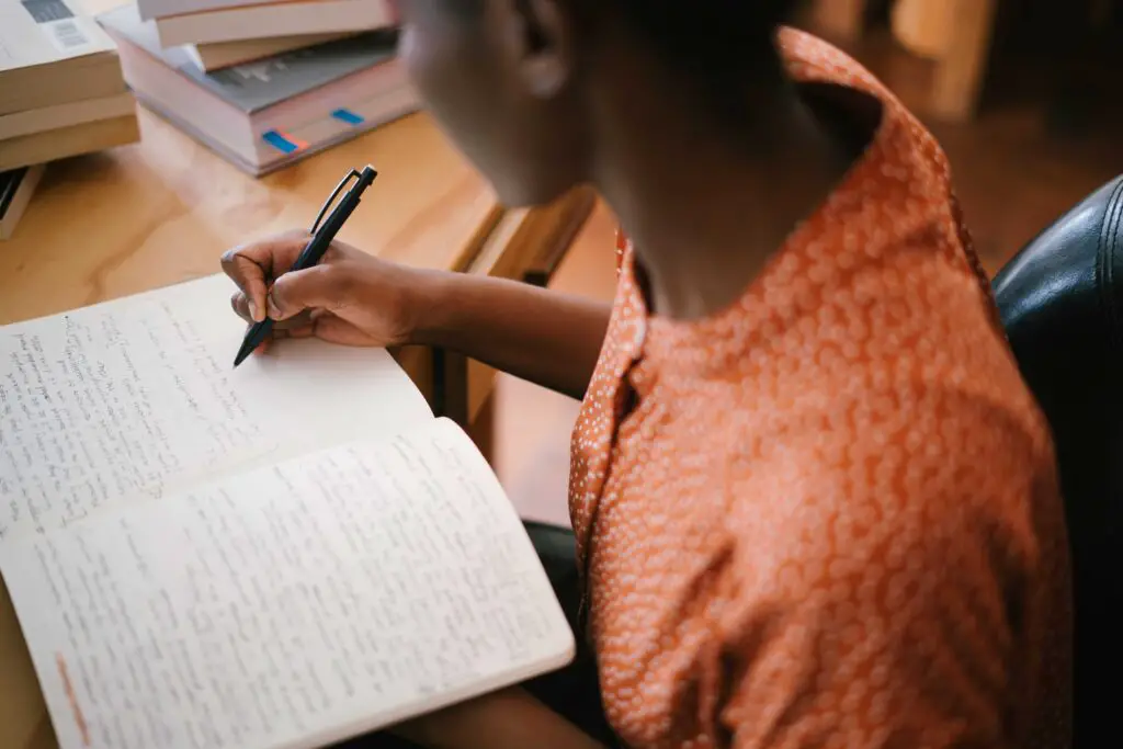 A black teenager in an orange shirt writing in an open notebook at a desk, maybe working on some of the listed language learning exercises.