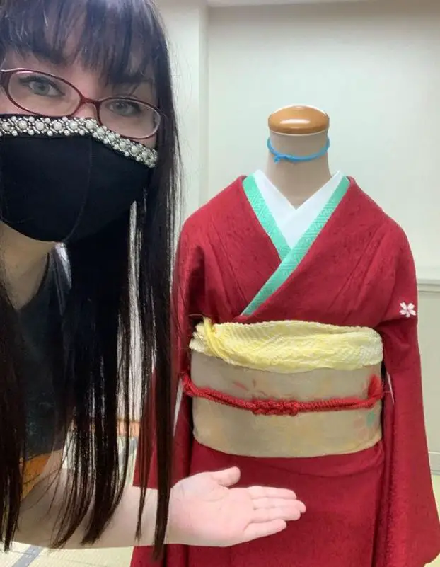 Jaenelle preparing for her kimono exam. Jaenelle wears a facemask and gestures to a mannequin wearing a red kimono and yellow obi.