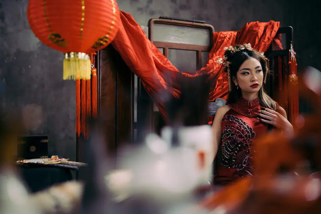 Featured image for Chinese-inspired fantasy booklist. A young woman from East Asia is dressed in a red dress and matching makeup, in a room with a red curtain and red laterns.