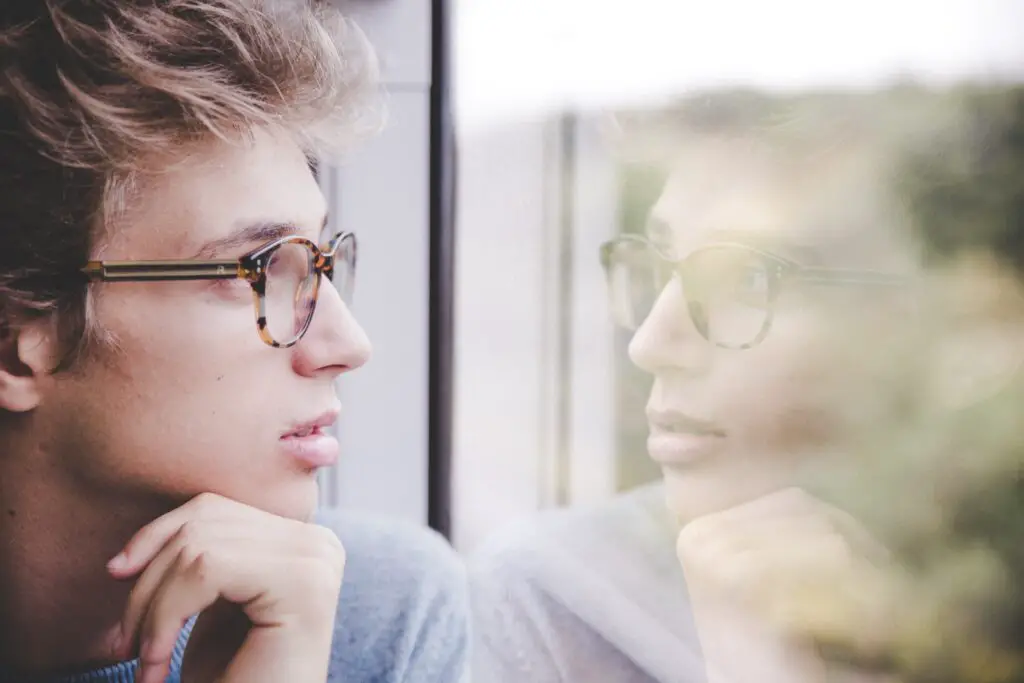 A young man with glasses and brown hair looking pensively at his reflection in a window. Featured image for foreign language anxiety article.