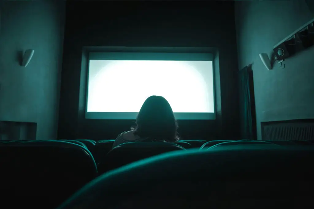 A woman sitting alone in front of a bright movie screen. featured image for Greek movies article.