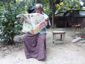 A photo of an elderly man sitting outside in a dhoti, reading a Bengali newspaper. Featured image for Bengali dialects article.